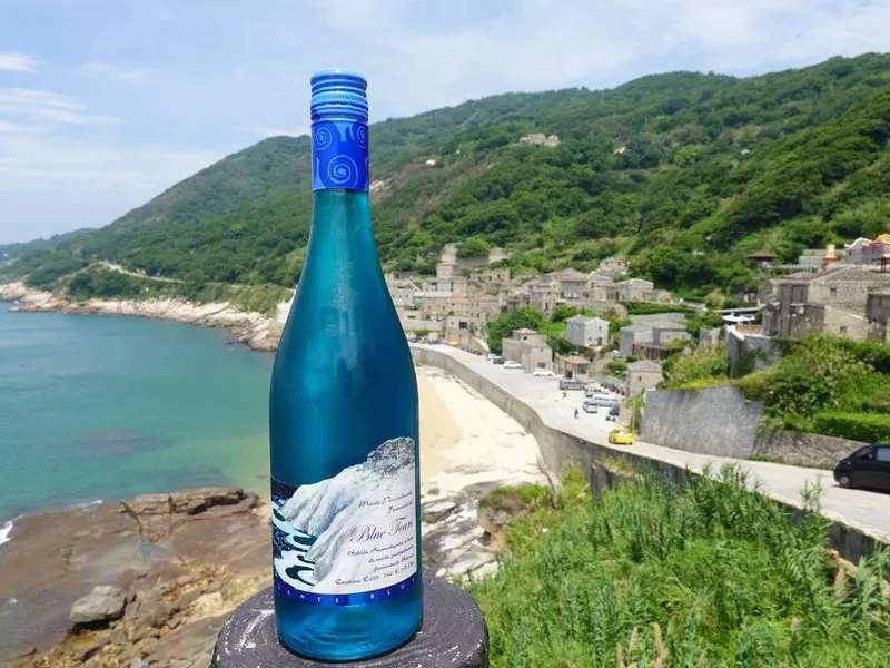 藍眼淚葡萄酒立在北竿芹壁海岸邊,後方是芹壁石屋聚落與海。